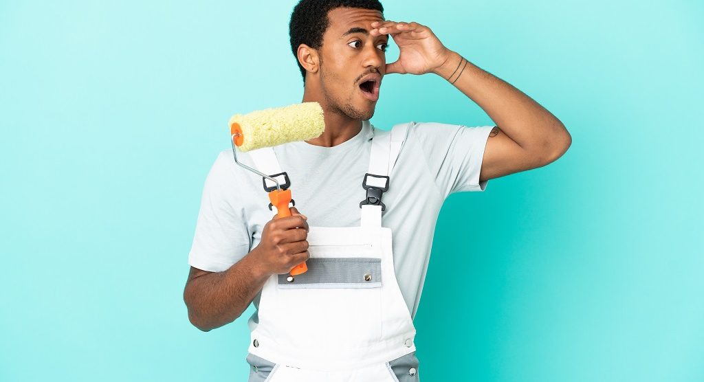 African American painter man over isolated blue background doing surprise gesture while looking to the side