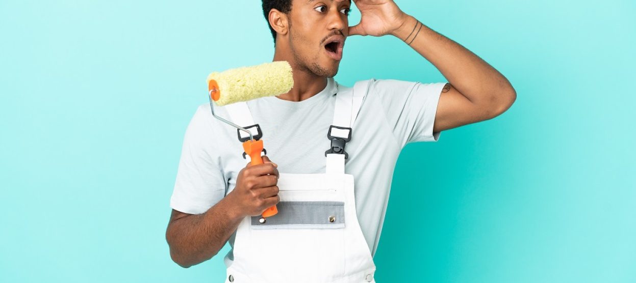 African American painter man over isolated blue background doing surprise gesture while looking to the side African American painter man over isolated blue background doing surprise gesture while looking to the side